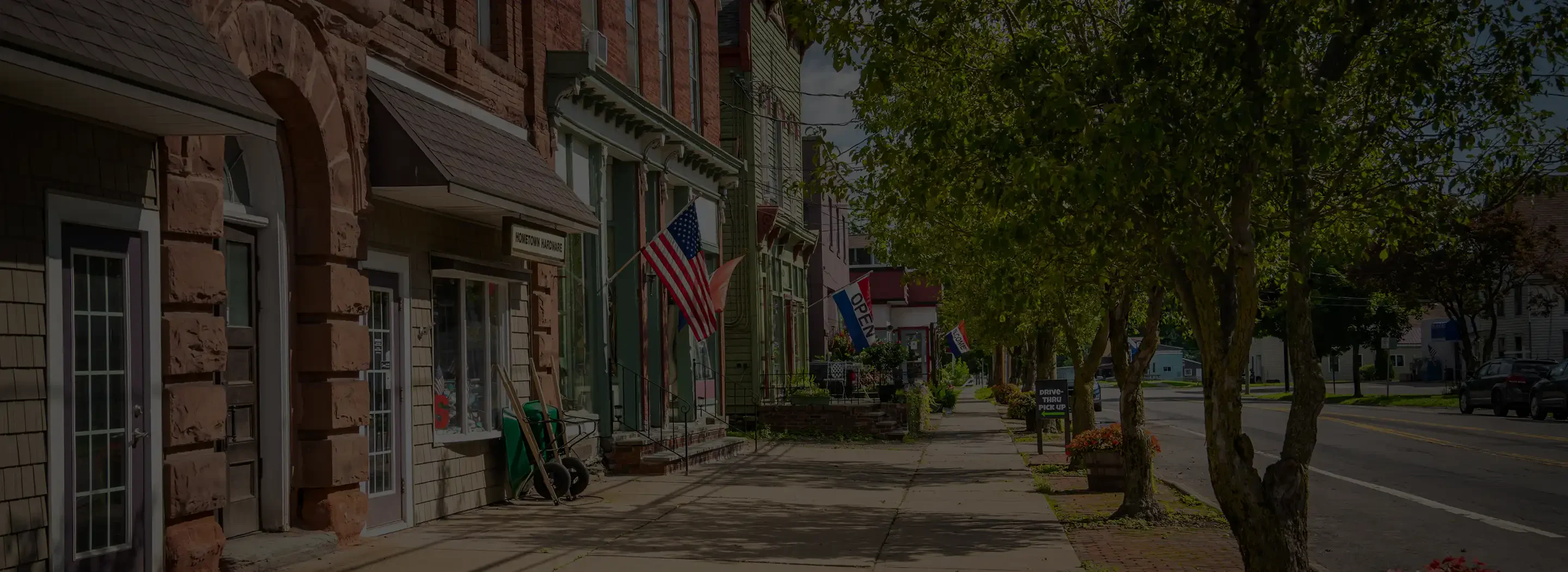 Charming street scene featuring colorful storefronts, American flags, and trees lining a peaceful sidewalk in a small town.