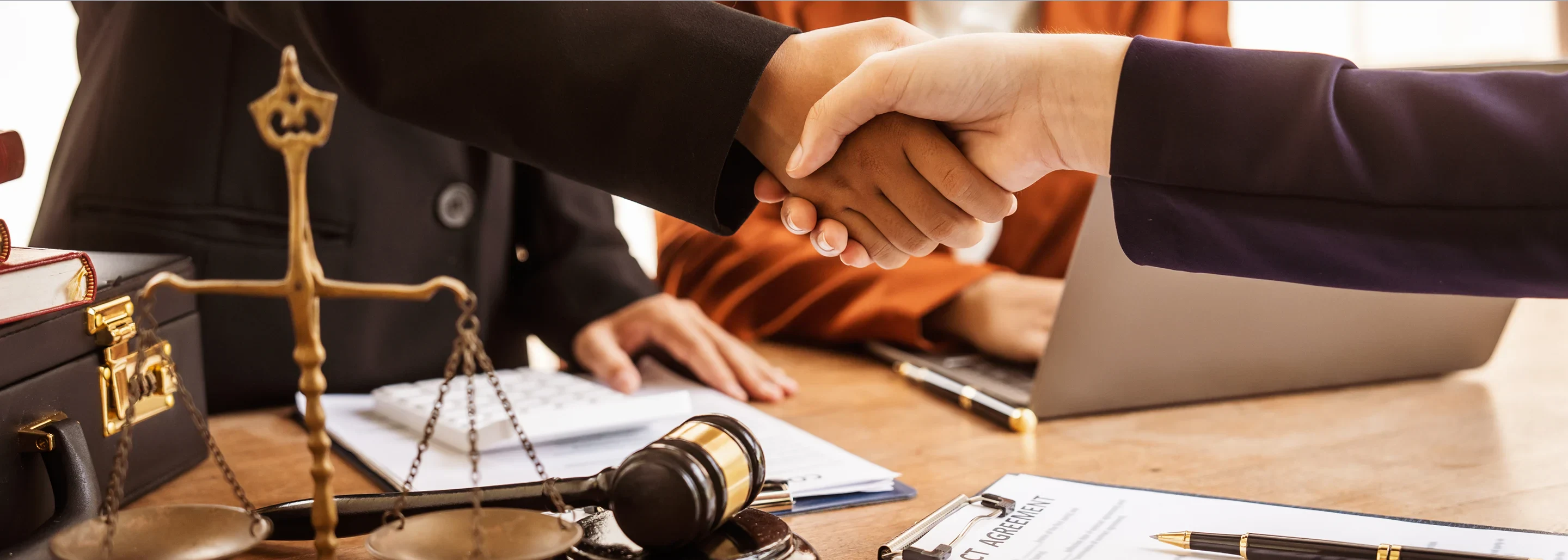 A close-up of two people shaking hands in a legal setting, with a gavel, scales of justice, and documents on a table.
